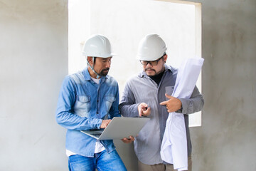 Portrait of two engineers or architects male, with safety helmets and laptop, collaborating on-site, reviewing digital blueprints and discussing the next steps in the construction project, men at work