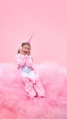 Child in pink unicorn costume sitting on pink fluffy surface, listening to music in headphones and singing against pink background. Concept of childhood, fantasy, dreams, imagination. Playful mood