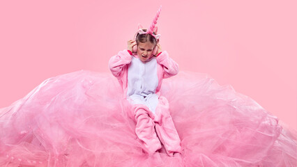 Child dressed in pink warm unicorn onesie, sitting on fluffy fabric and listening to music with headphones against pink background. Concept of childhood, fantasy, dreams, imagination, emotions © master1305