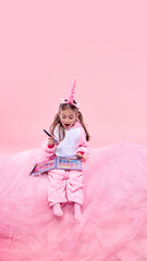 Girl in pink unicorn onesie sitting with makeup palette, holding brush, expressing excitement and pure joy against pink studio background. Concept of childhood, fantasy, dreams, imagination
