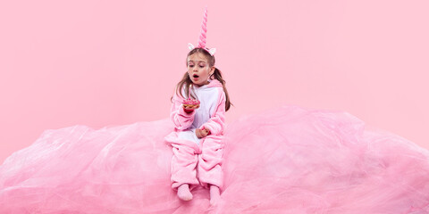 Child girl in pink unicorn onesie holding pink donut and blowing lit candle against pink background. Happy birthday celebration. Concept of childhood, fantasy, dreams, imagination © master1305