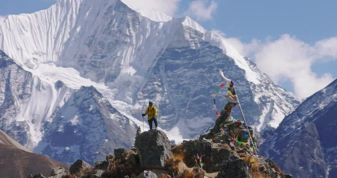 Drone shot with telephoto zoom of a male hiker reaching the summit in Nepal Mountain. Surrounded by high-altitude snow-capped Himalayas including Everest and Langtang, adorned with prayer flags.