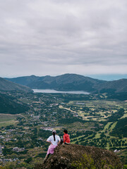 Naklejka premium A Woman and a Child Enjoy the Panoramic Scenery of a Landscape with Hills, a Lake and a Town from a Mountain Perch; Two People Sit on a Rocky Outcrop, Gazing at the Breathtaking View of a Valley