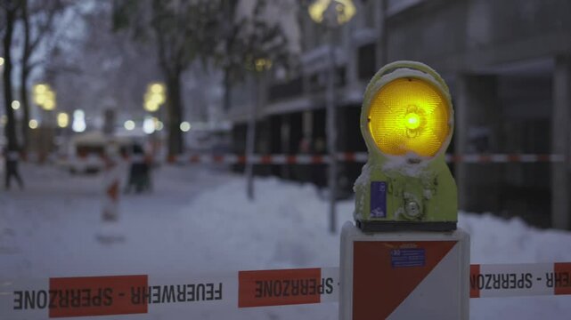 Street in Munich, Germany, is closed due to heavy snowfall. A flashing warning light and barrier tape mark danger zone, alerting pedestrians to risk of falling snow. Safety measures are in place. 