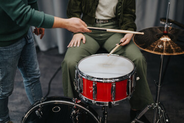 Two individuals practicing drumming techniques in a studio setting using a vibrant red drum kit. The image captures a close-up atmosphere of a hands-on music teaching experience.