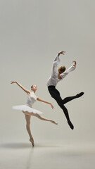 Naklejka premium Poised ballerina on pointe with male partner leaping beside, showcasing contrast between lightness and strength against white studio background. Concept of classic art and beauty, visual harmony.