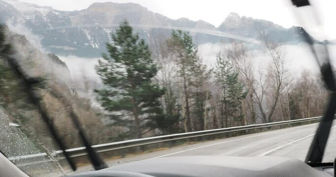 Navigating treacherous mountain road, windshield wipers clearing rain and snow while low clouds obscure dramatic alpine landscape during challenging winter driving conditions