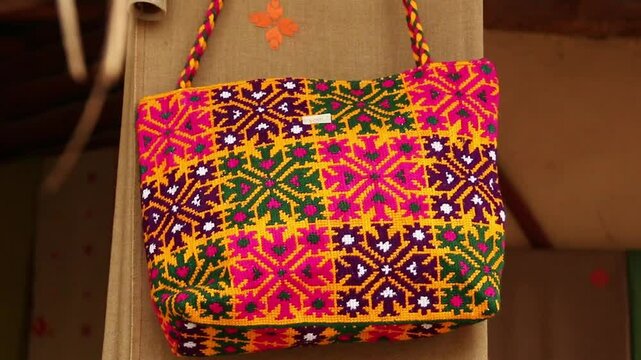 Colorful embroidered textile bag with geometric and floral patterns, orange and purple tones, fringed edges. 
