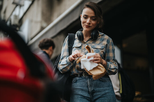A young woman with headphones enjoys a snack while walking on a busy city street, capturing a moment of urban lifestyle and casual fashion.