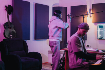 Two musicians work together in a contemporary recording studio. One sings into a microphone while the other plays a piano keyboard. The setting includes guitars and recording equipment.