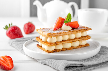 French dessert millefeuille with vanilla cream and fresh strawberries on a white plate on a white wooden background.