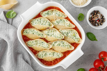 Conchiglioni pasta stuffed ricotta cheese and spinach with tomato sauce in a white baking dish on a gray concrete background.