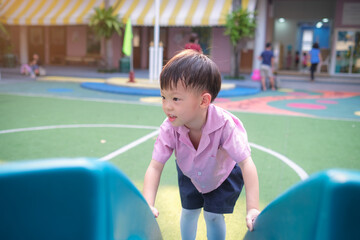A cheerful 4-year-old Asian kindergarten boy enjoys playing on a slide at school playground. With a bright smile, he experiences fun, adventure, and active play in an outdoor learning environment