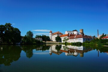 Obraz premium A serene view of Jindřichův Hradec Castle in the Czech Republic, reflecting beautifully in the calm waters of the pond. The castle's white walls and red rooftops stand out against the clear blue sky.