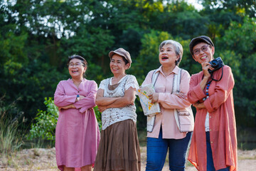 Asian senior women standing together in nature, laughing with happiness, celebrating their retirement lifestyle. Holding a map and camera, they share a joyful moment on a relaxing outdoor trip.