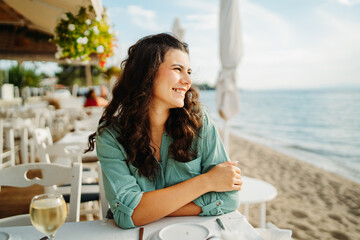 Young caucasian woman eat greek salad and drink wine in restaurant by the sea	
