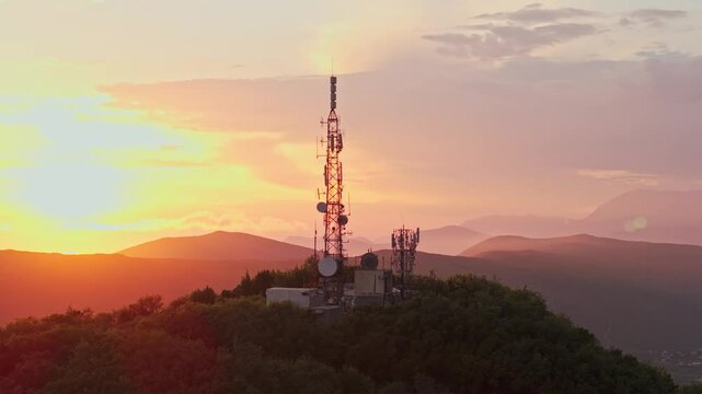 Aerial drone footage of a telecommunications tower on a green hill at sunset. Antennas and satellite dishes highlight modern connectivity, with mountains in the background. Perfect for themes of 5G, n