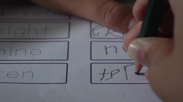 angled shot of a child's hand holding a green pencil and writing on a worksheet. The worksheet features boxes with words printed in a simple, educational font, suggesting a learning activity.
