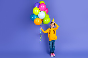 Fototapeta premium Adorable preteen girl in casual yellow sweatshirt with colorful balloons, celebrating with happy expression over purple background