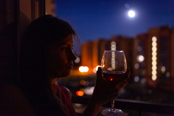 A girl drinks from a glass near the window in the evening looking at the city lights