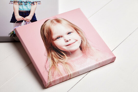 Close-up of a gallery-wrapped canvas print featuring a smiling young blonde girl on pink background, displayed on a white wooden surface, perfect as personalized home decor or gift