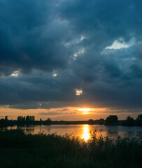 beautiful sunset, warm rays break through dark clouds over the lake and the shore with reeds