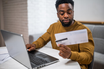 Focused young African-American man using laptop and credit card to pay bills online, managing his...