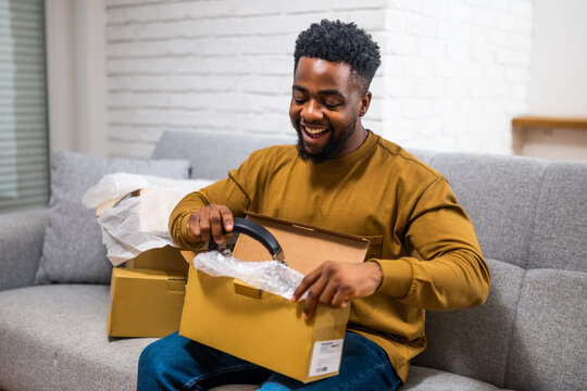 Young black man sitting comfortably in living room and unboxing package with joy after receiving online shopping delivery. Representation of customer satisfaction with home shopping.