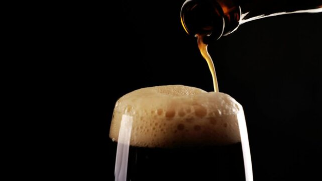 extreme closeup of slow motion pouring dark beer from beer bottle on whitecap, head of beer illuminated and isolated on black background, motion design concept