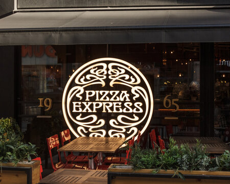 Bradford, Yorkshire, UK 2-24-2025 Illuminated logo of Pizza Express lights up a table and chair outside one of their more than 500 pizza restaurants