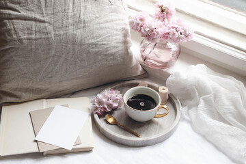 Spring still life scene. Blank greeting card invitation mockup. Cup of coffee on marble tray, old books. Feminine styled photo. Floral composition. Pink sakura cherry tree blossoms, table near window.