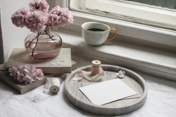 Spring still life scene. Greeting card mockup, marble tray, cup of coffee, old books. Vintage feminine styled photo. Floral composition. Pink sakura, cherry tree blossoms on table near window.