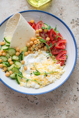 White and blue plate with yogurt, cucumber, tomatoes, chickpeas and grated egg, vertical shot on a beige granite background, high angle view