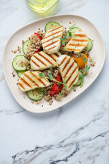 Beige plate with quinoa, grilled halloumi cheese and vegetables, vertical shot on a white granite background, high angle view
