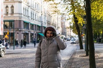 Fototapeta premium Cropped view of young adult tired night shift worker man walking along Helsinki's city street with classical European buildings & trees wearing grey hooded puffer jacket and backpack with hand on head