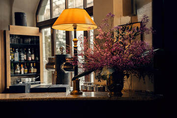A warmly lit bar interior with an elegant table lamp, a lush bouquet of pink flowers, and a stylish bar counter. Perfect for hospitality, luxury, and lifestyle imagery.