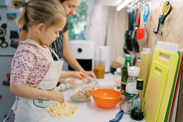 Little girl puts toppings on rolled out pizza dough while standing with her mom at the kitchen table
