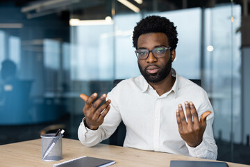 An African American man wearing glasses seriously communicates during a video call from his modern office, gesturing with his hands