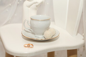 Elegant cup with sweets and wedding rings on white background