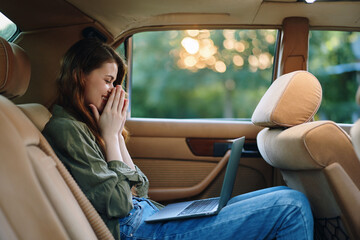 A stressed young woman in casual attire sitting in the backseat of a car, working on a laptop, surrounded by green nature during sunset, reflecting on challenges.