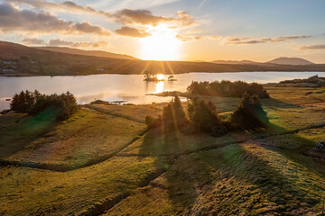 Warm sun flares cast a golden glow over a tranquil Connemara lake, enhancing the ethereal beauty of the landscape.