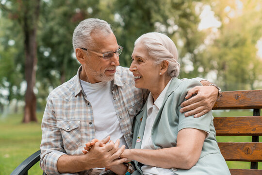 Smiling senior couple embracing and holding hands while resting on wooden bench in park looking to each others. Support elderly love active seniors future life concept
