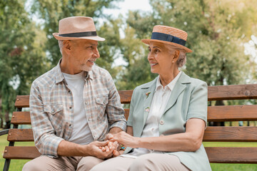 Obraz premium Smiling senior couple in hats embracing and holding hands while resting on wooden bench in park looking to each others. Support elderly love active seniors future life concept