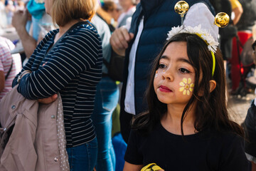 A young girl with a yellow flower painted on her face and a bee headband is standing in a crowd