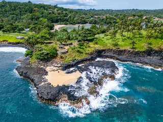 Boca de Inferno, Sao Tome, featuring black volcanic rock formations, turquoise ocean waves, and a lush green hillside covered with tall, swaying palm trees under a clear blue sky.