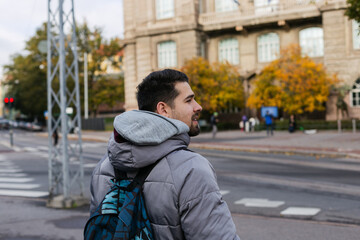 Rear view of a young adult bearded man walking on Helsinki's streets in casual grey puffer jacket with blue backpack turning their head. Urban cityscape background © Anna