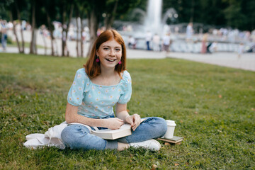 Young happy teenage girl with freckles reading a book sitting on the grass in a public park