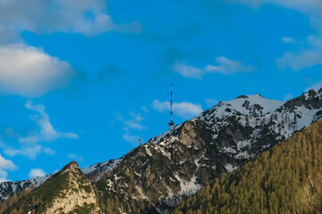 Iconic transmitter tower atop snow-capped Dobratsch mountain stands tall against clear blue sky. Jagged rocks and patches of evergreen forest cling to slopes, revealing rugged terrain of Gailtal Alps