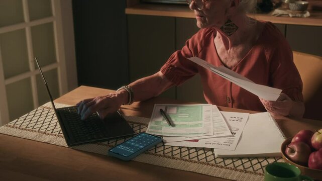 Focused elderly lady comparing costs of bills with previous month using laptop in kitchen after increasing of tariffs on utility services