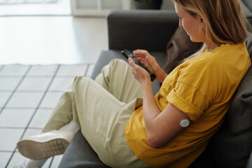 Woman with diabetes sitting on couch at home with smartphone and CGM sensor on arm